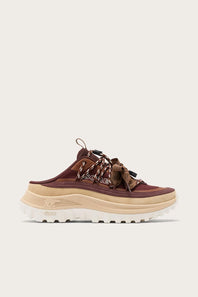 A single low-top sneaker shown in side profile against a light off-white background. The shoe features a dark brown suede upper with a matching brown lace, a thick beige midsole, and a white lug sole.