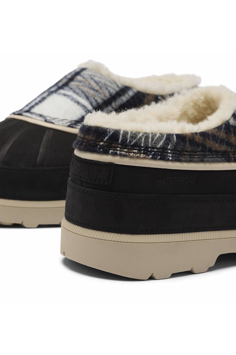Close-up of a pair of slip-on mule shoes with a black suede-like side, a plaid patterned upper, cream shearling lining, and a thick beige sole, shown on a white background.