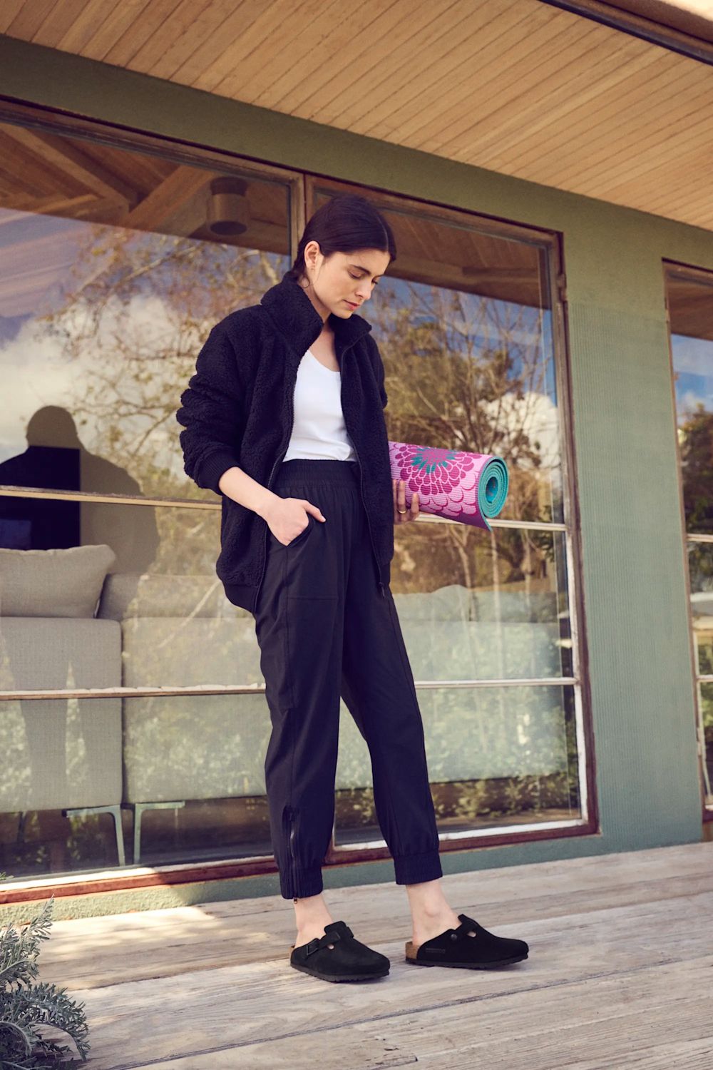 A woman stands on a wooden deck in front of a modern house, wearing a black fleece jacket, black cropped pants, and black slip-on shoes. She has a white tank top underneath.