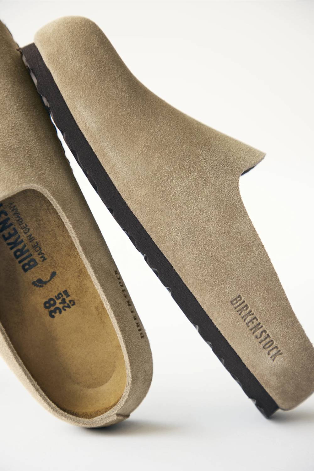 A pair of Birkenstock slip-on clogs in natural suede, showing the side profile and the interior footbed with the brand name 'BIRKENSTOCK' and size '38' printed on it.