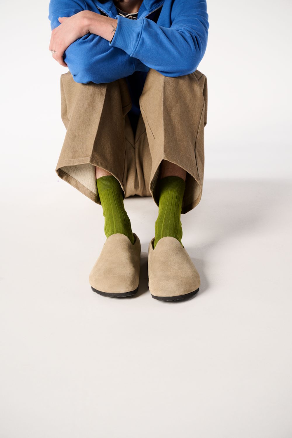 A person seated against a plain light background, wearing a bright blue top, loose tan pants, bright green socks, and tan slip-on shoes.