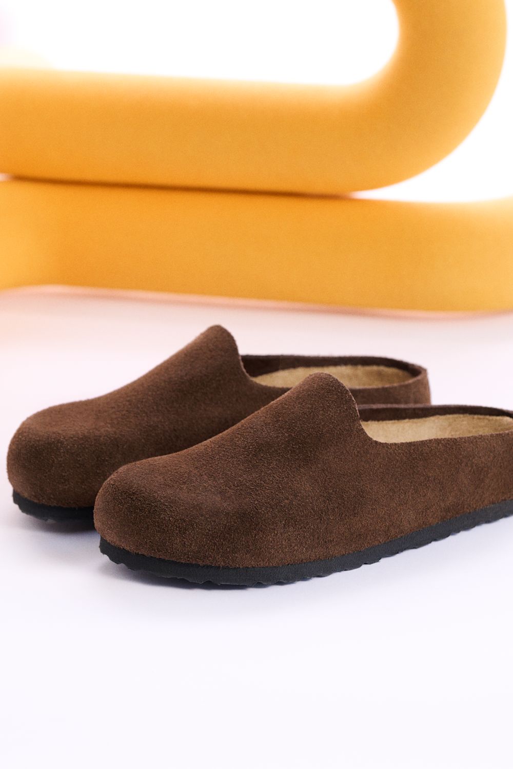 A pair of dark brown suede slip-on mule slippers with black soles, displayed on a white surface with a yellow abstract background prop.