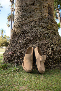A pair of tan leather penny loafers resting on green grass in front of a large tree trunk outdoors.