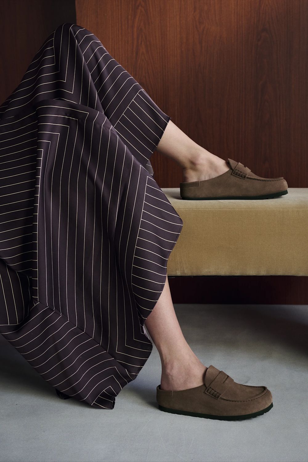A person seated indoors wearing brown slip-on loafers, with one shoe on the foot and the other resting on a bench. A dark pinstriped garment is draped over the leg, and the setting includes a wooden wall and gray floor.