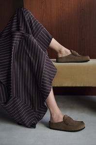 A person seated indoors wearing brown slip-on loafers, with one shoe on the foot and the other resting on a bench. A dark pinstriped garment is draped over the leg, and the setting includes a wooden wall and gray floor.