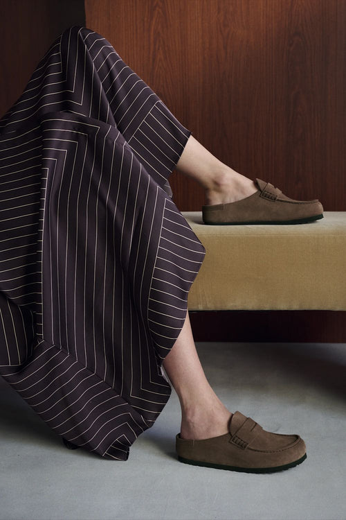 A person seated indoors wearing brown slip-on loafers, with one shoe on the foot and the other resting on a bench. A dark pinstriped garment is draped over the leg, and the setting includes a wooden wall and gray floor.
