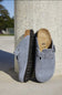 A pair of light blue suede clogs with black rubber soles, leaning against a concrete pillar outdoors.