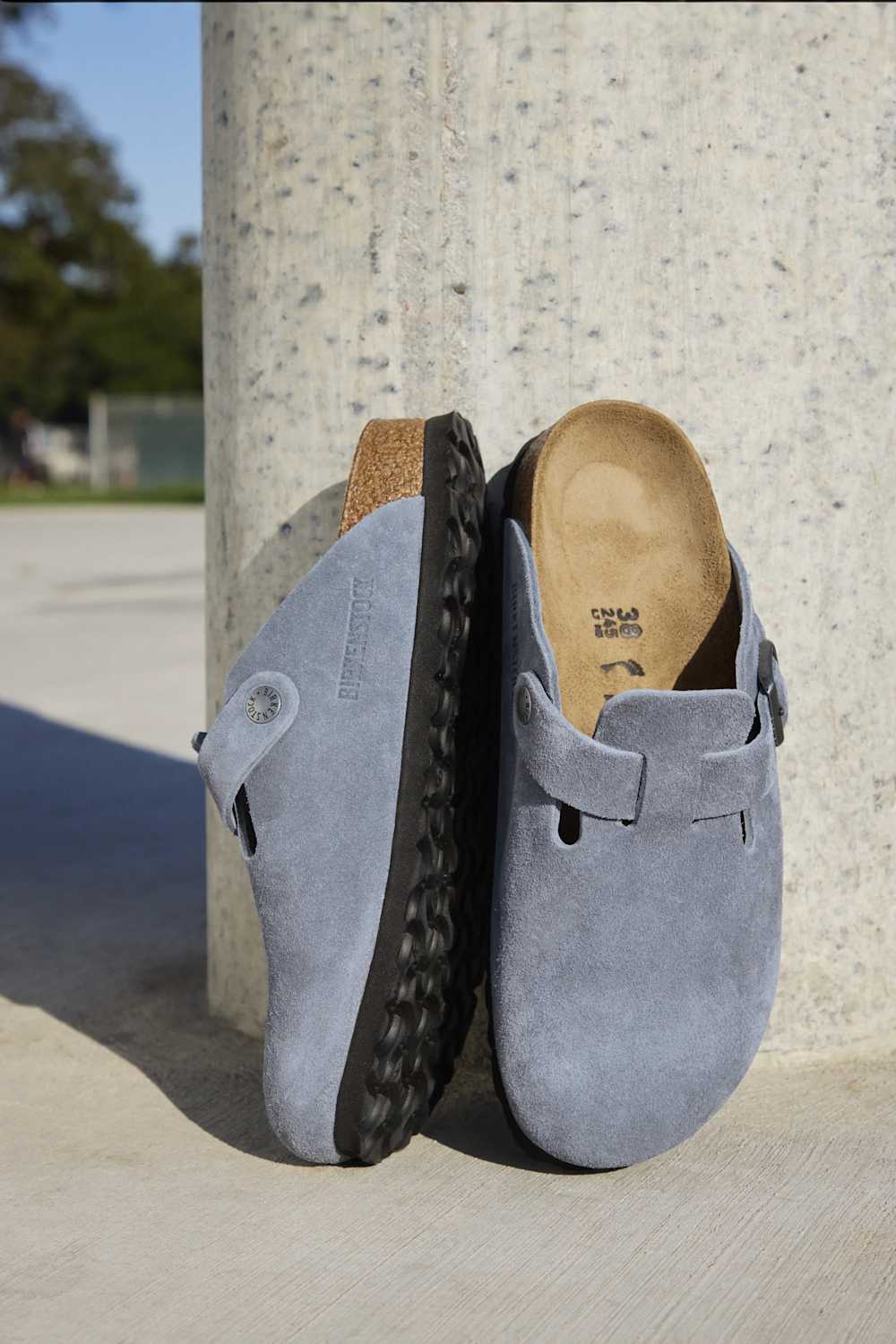 A pair of light blue suede clogs with black rubber soles, leaning against a concrete pillar outdoors.