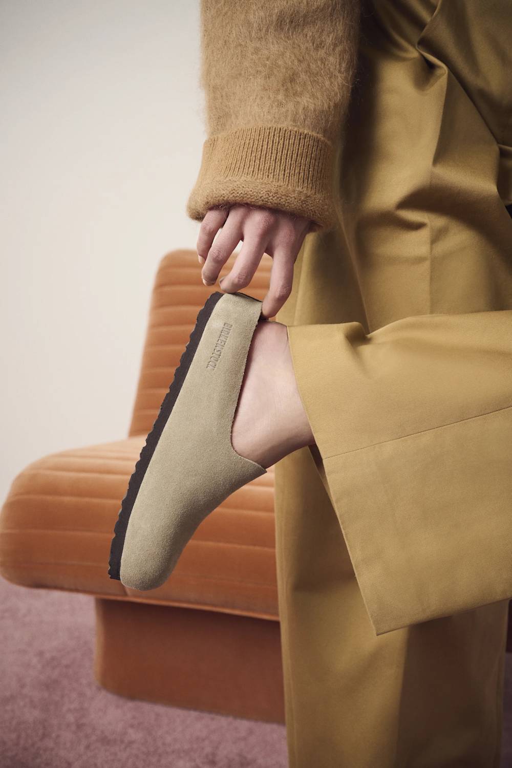 A person wearing a camel-colored knit sweater and matching wide-leg trousers lifts a tan slip-on shoe with a black sole, posed in front of a modern orange chair.