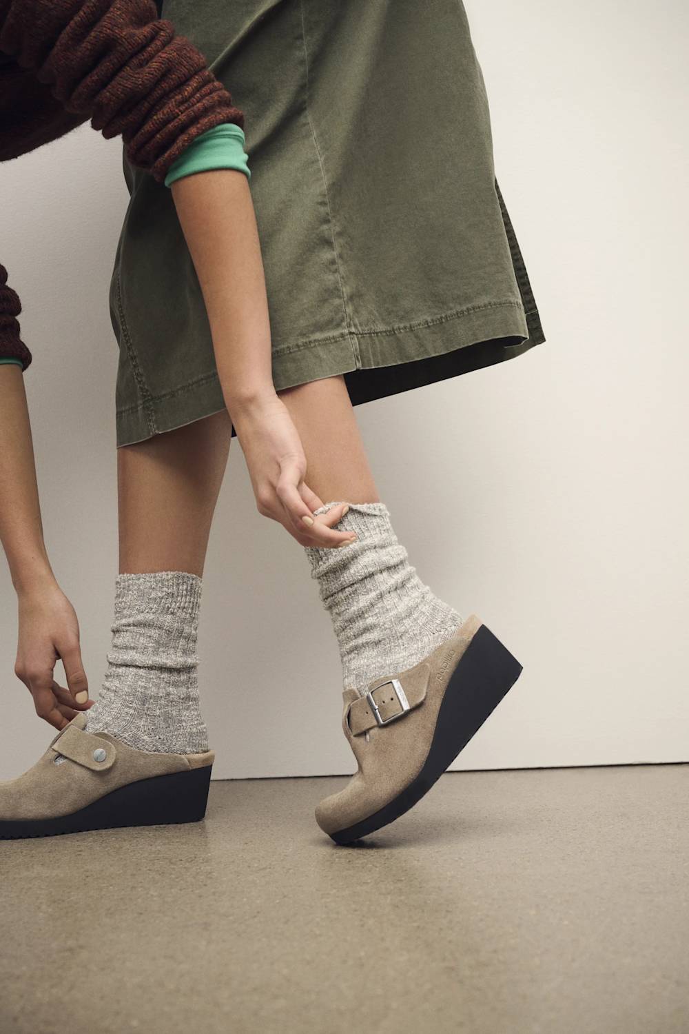 A person wearing taupe wedge clogs with buckle straps and light gray heathered crew socks, shown from the waist down in an indoor setting.