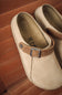 A pair of beige suede clogs with brown leather straps and silver-tone metal buckles, resting on a brown leather seat.