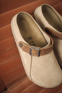 A pair of beige suede clogs with brown leather straps and silver-tone metal buckles, resting on a brown leather seat.