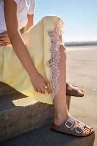 A person sitting outdoors wearing a pale yellow dress with a sheer white lace panel down the side and brown slide sandals with large silver buckles.