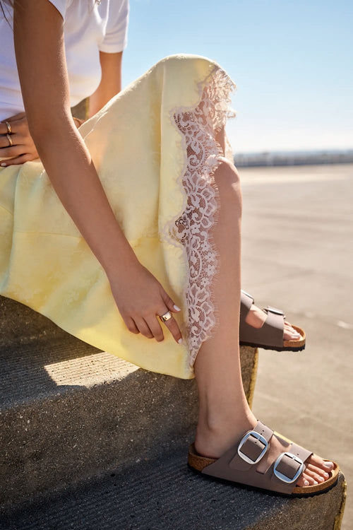 A person sitting outdoors wearing a pale yellow dress with a sheer white lace panel down the side and brown slide sandals with large silver buckles.