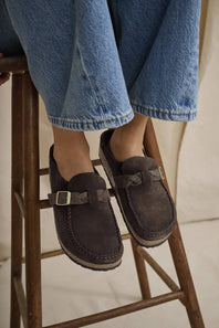 A person wearing blue jeans sits on a wooden stool, showing a pair of dark brown suede slip-on shoes with woven strap details and small silver buckle accents.