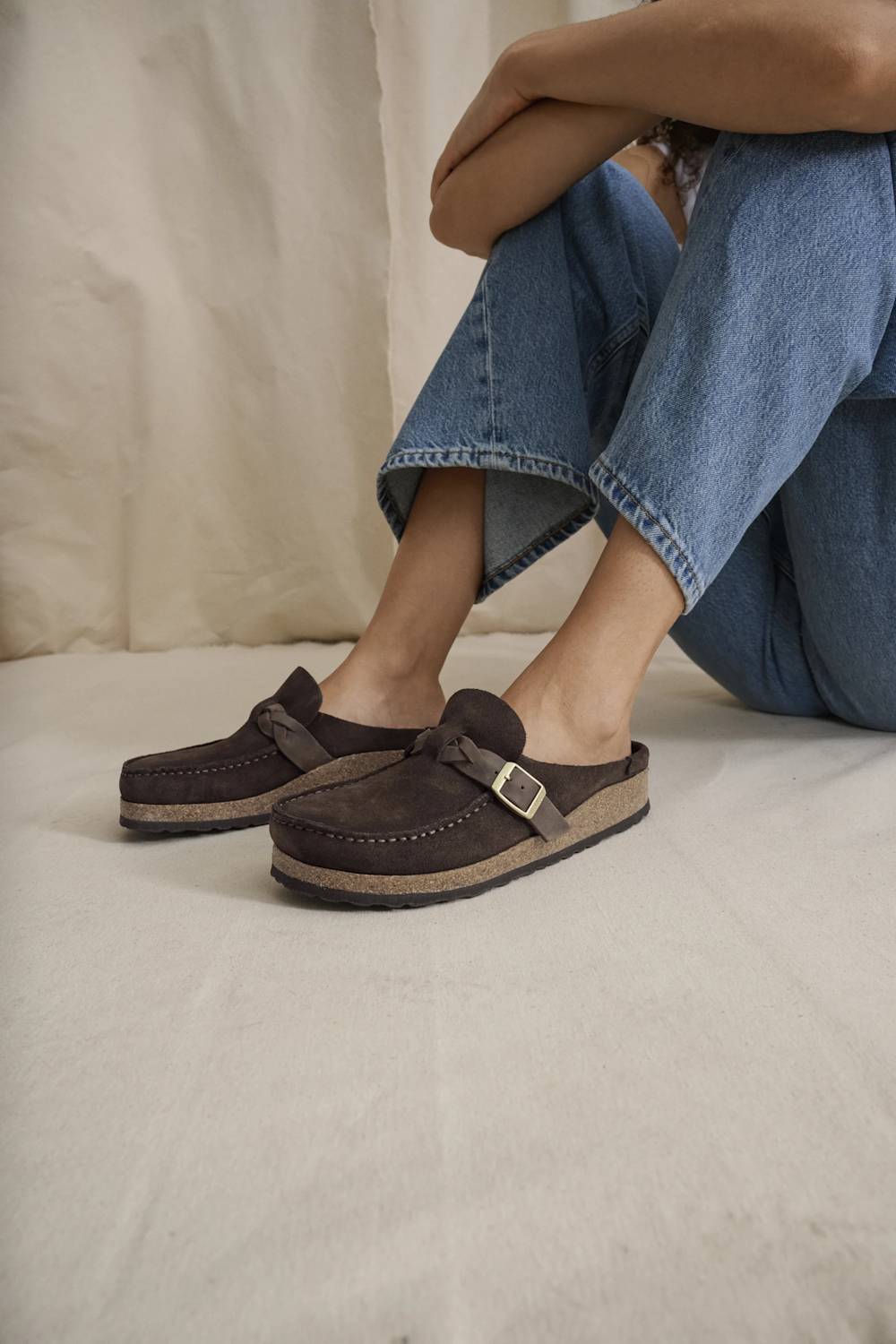 A person seated on a light fabric surface wearing dark brown slip-on mule clogs with a small metal buckle strap and light tan soles, paired with blue jeans.