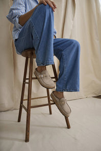 Person wearing blue jeans and beige loafers sitting on a wooden stool against a neutral background