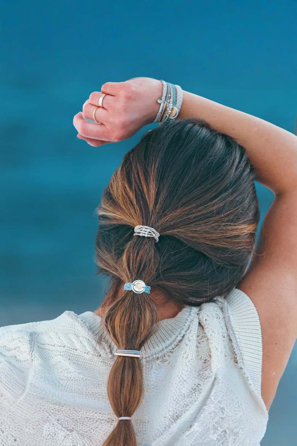 A woman viewed from behind with her hair in a low ponytail, wearing several silver hair ties and bracelets against a blue outdoor background.