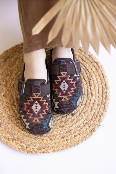 Person wearing patterned clogs on a woven mat with a plant leaf in the background