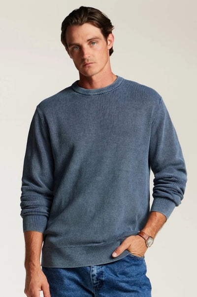 Man standing in front of a plain studio background wearing a slate blue long sleeve sweater with the sleeve rolled up to mid forearm with blue denim jeans