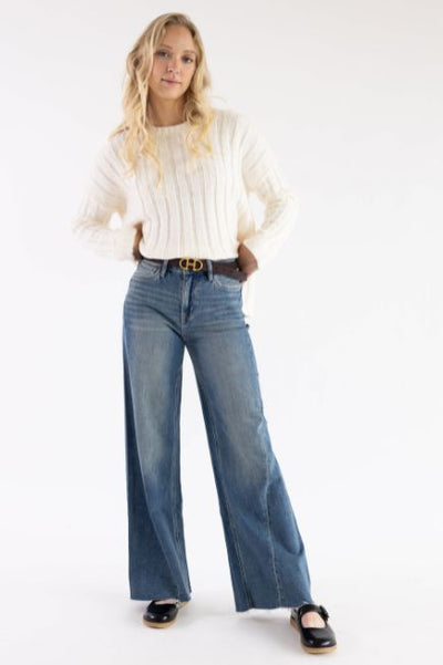 A woman standing in front of a basic studio background wearing a white ribbed sweater and denim jeans styled with a belt and black Mary Jane shoes