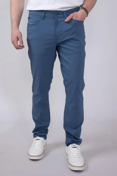 Person shown from the waist down wearing slate blue dress pants with a white shirt and white sneakers while standing in front of a plain studio background