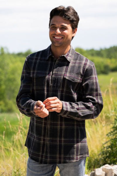 Man standing in a grassy outdoor area wearing a dark grey and burgundy plaid flannel shirt and denim jeans