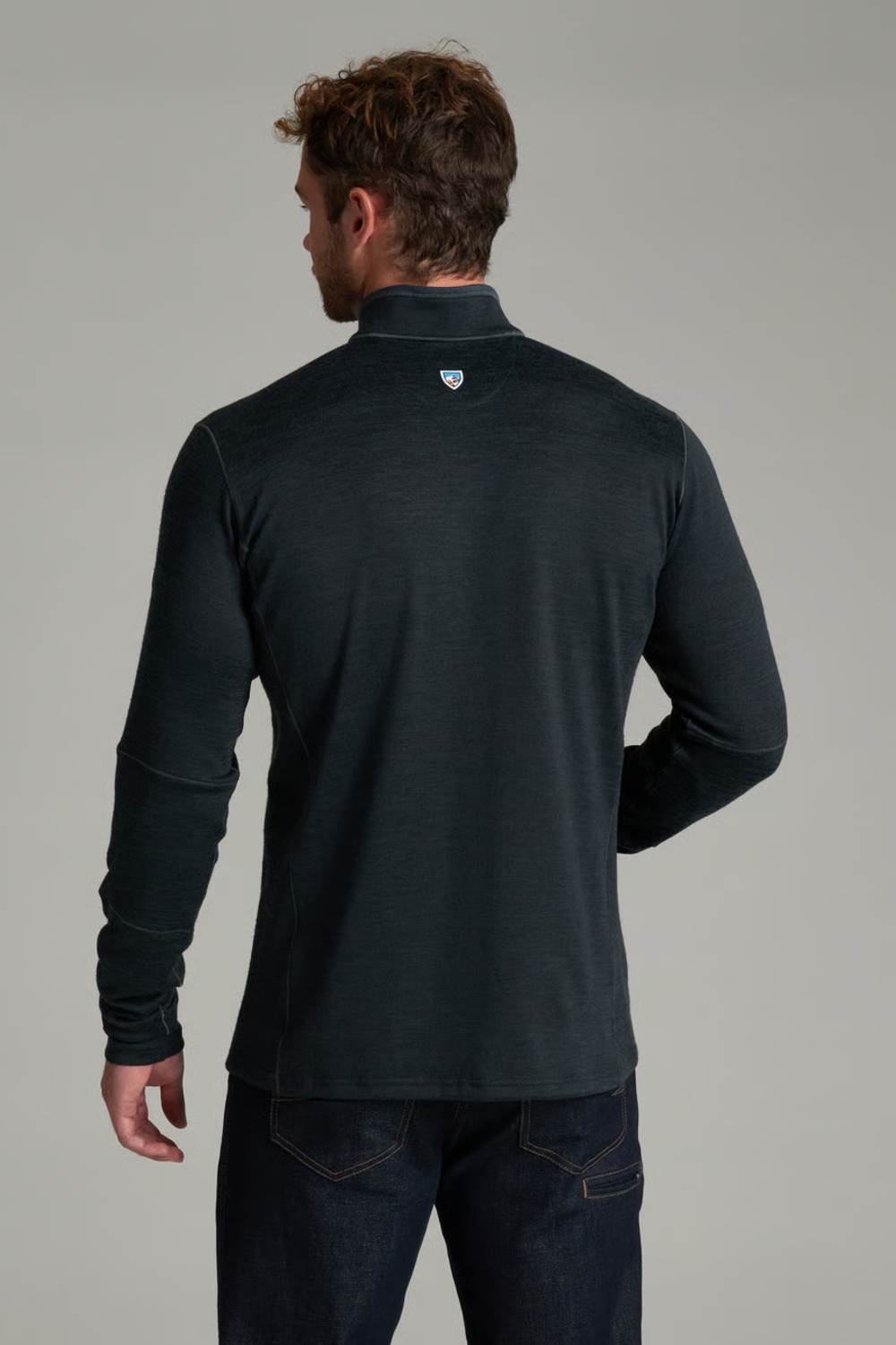 A man viewed from the back wearing a dark charcoal gray long-sleeve quarter-zip pullover and dark blue jeans, against a solid light gray studio background.