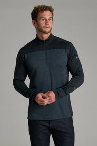 A man wearing a dark charcoal and black quarter-zip long-sleeve athletic shirt, paired with dark jeans, standing against a solid gray studio background.