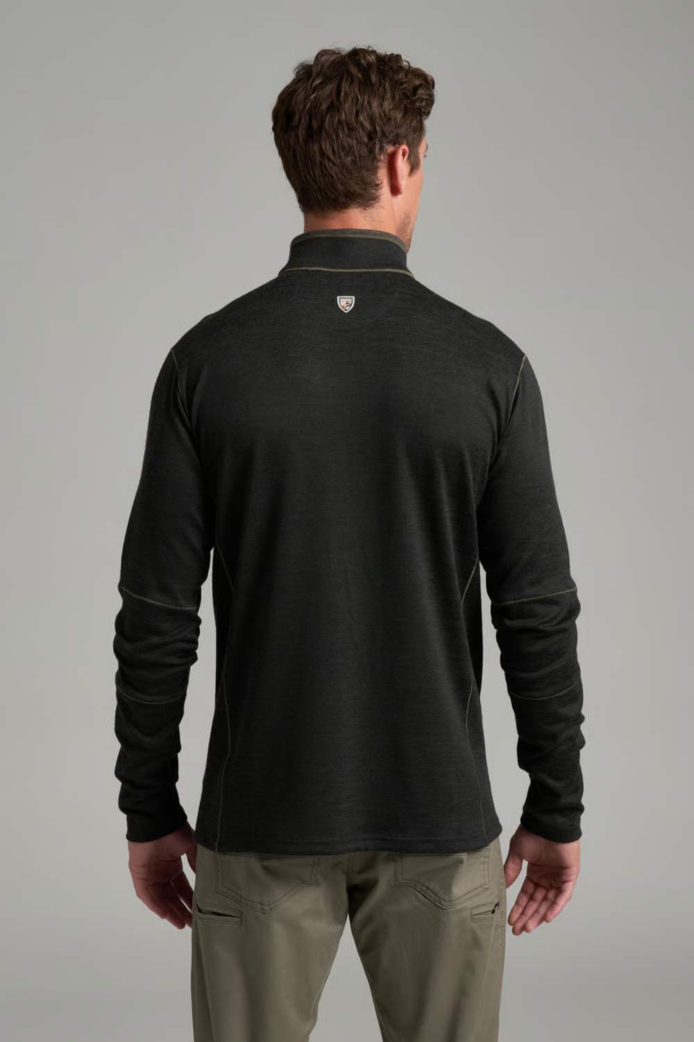 A man viewed from behind wearing a dark charcoal gray long-sleeve quarter-zip pullover with a small shield logo on the upper back, paired with khaki pants, against a neutral gray studio background.