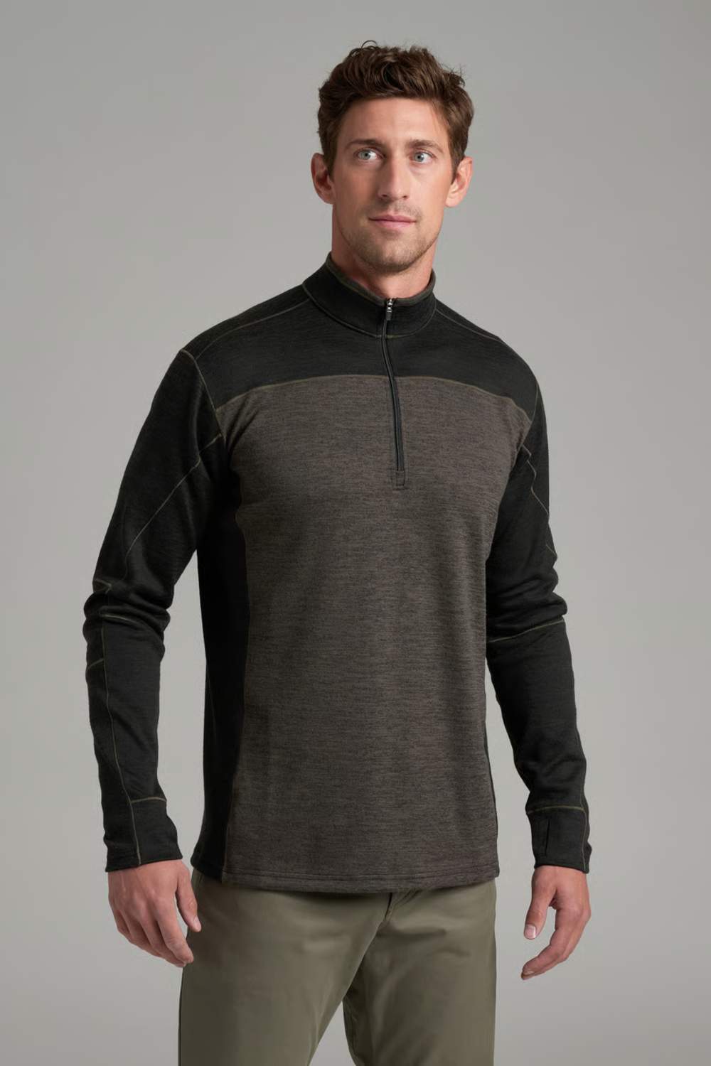 A man wearing a dark gray and black color-blocked quarter-zip long-sleeve athletic shirt, paired with olive green pants, against a solid gray studio background.