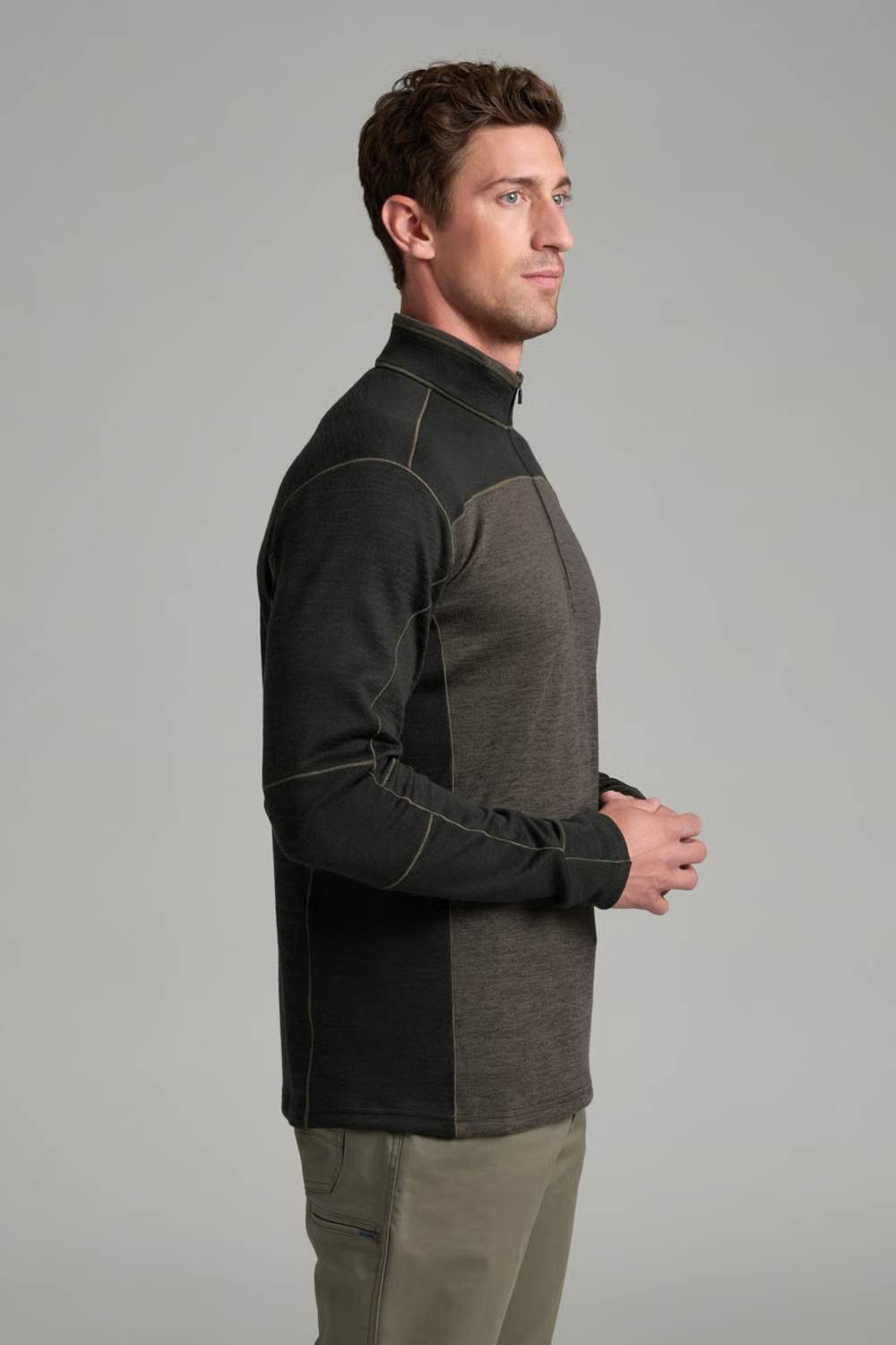 A man in side profile wearing a color-blocked quarter-zip fleece pullover with dark charcoal and heather gray panels, paired with khaki pants, against a solid gray studio background.