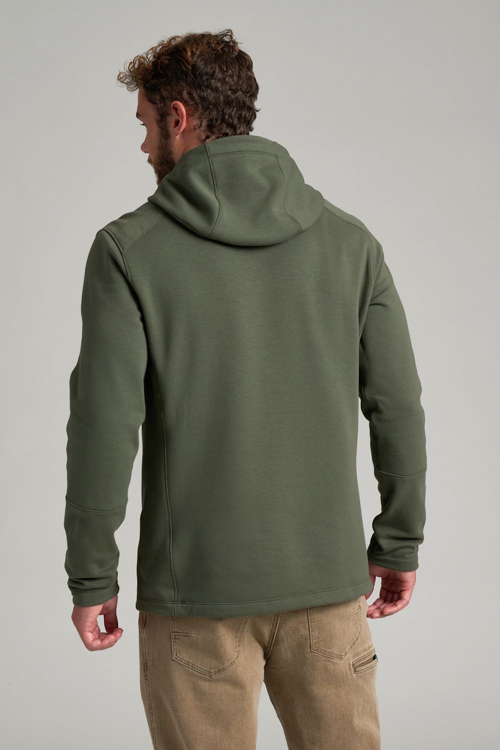A man wearing an olive green hooded sweatshirt, shown from the back, paired with khaki pants, against a solid light gray studio background.