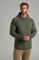 A man wearing an olive green hooded sweatshirt and tan pants, standing against a neutral gray studio background.