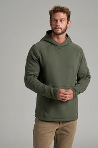 A man wearing an olive green hooded sweatshirt and tan pants, standing against a neutral gray studio background.
