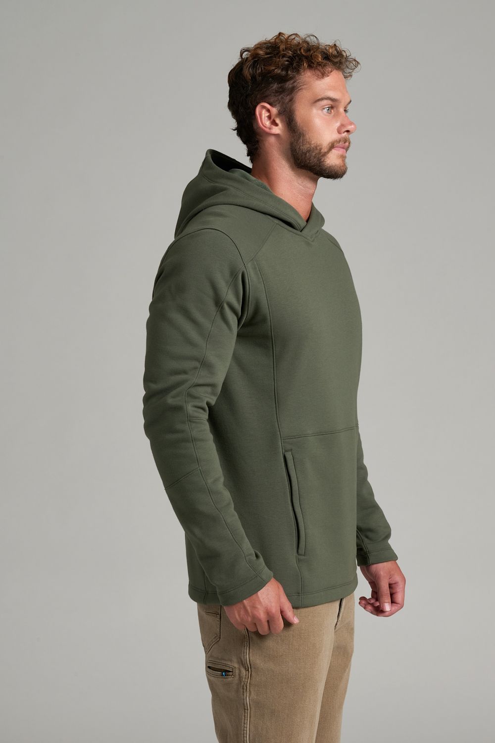 A man wearing an olive green pullover hoodie with a kangaroo pocket, shown from a side profile against a neutral gray studio background.