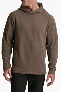 A man wearing a brown hooded sweatshirt with a kangaroo pocket, paired with black pants, shown from the chin down against a light gray background.