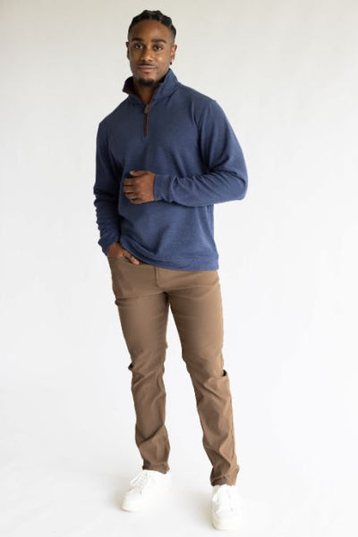 A man wearing a navy blue quarter zip sweatshirt with light brown pants and white sneakers standing in front of a basic studio background