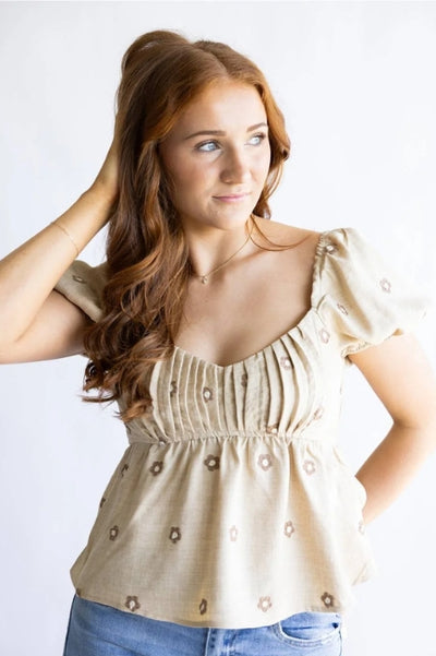 Woman wearing a V-neck light tan with embroidered brown and white flowers and blue denim jeans standing in front of a plain studio background