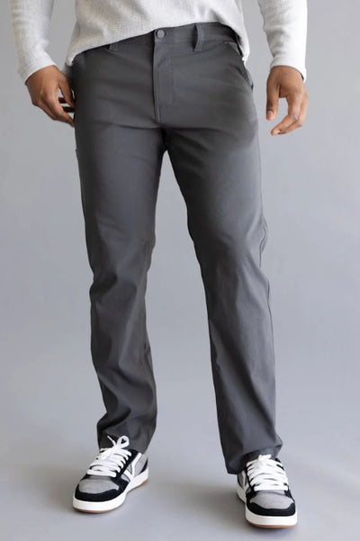 Person viewed from the waist down wearing dark grey dress pants with a long sleeve white shirt and black, white, and grey sneakers on a grey studio background