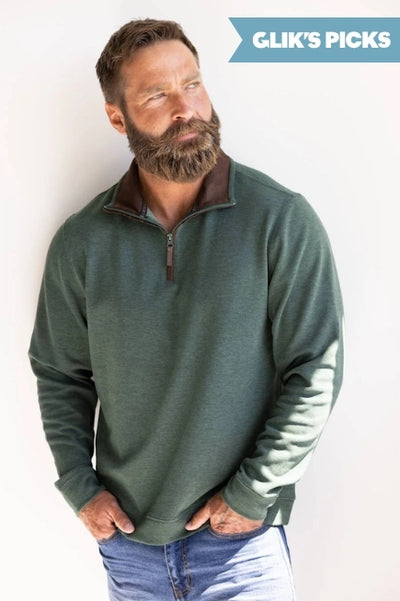 A man wearing a green quarter zip sweatshirt with denim jeans standing against a plain wall and a blue flag badge in the upper right corner that read GLIK'S PICKS