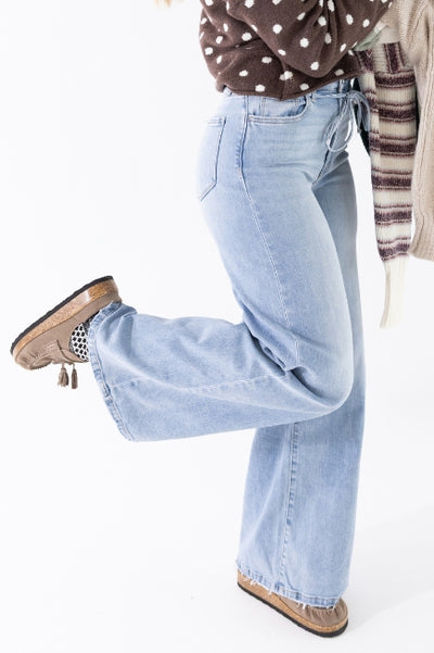A woman kicking her leg back wearing light wash denim jeans with a brown sweater with white polka dot and Moccasin style mules on a white background