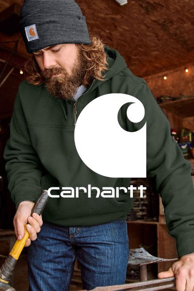 Image of a man wearing a grey Carhartt beanie and dark green quarter zip sweatshirt in a workshop and hammer in hand. The Carhartt logo in white is overtop in the middle of the image