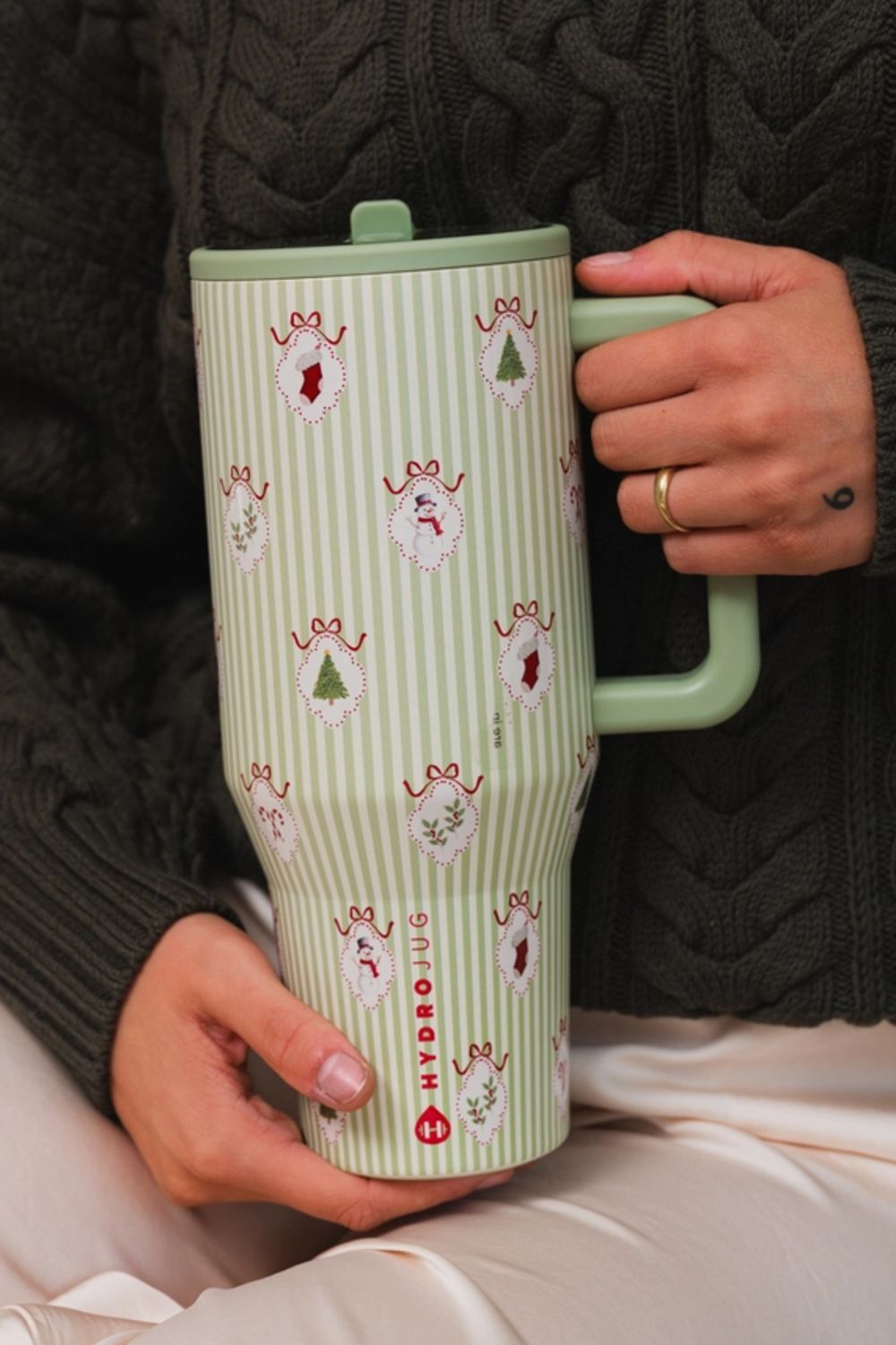 A person holding a HydroJug travel tumbler with a light green and white striped pattern decorated with small holiday motifs, wearing a dark cable-knit sweater.