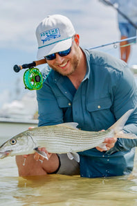 A smiling man in a blue button-up shirt, white cap, and sunglasses holds a striped bass fish while standing in shallow water on a boat. A green fly reel and fishing rod are visible behind him.