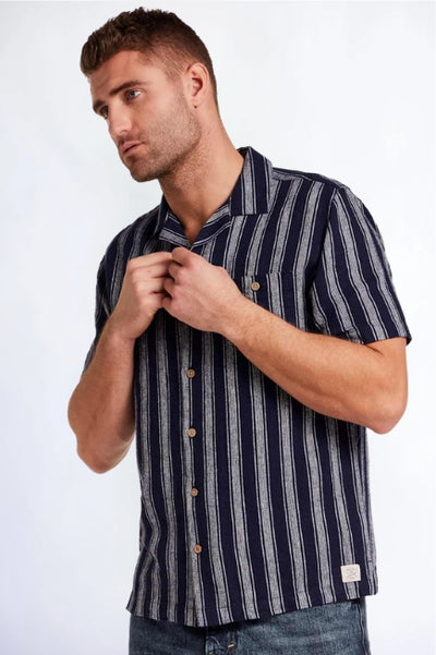 Man wearing a navy and grey vertical striped button up shirt and blue denim jeans standing in front of a studio background