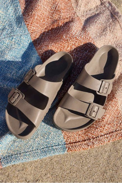 A pair of taupe double-buckle slide sandals rests on a multicolored woven blanket spread over sand, lit by natural sunlight.