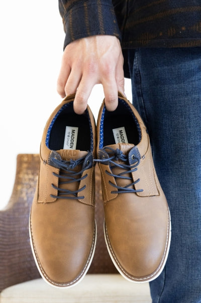 Zoomed in photo of a person holding a pair of brown dress shoes with the MADDEN brand logo on the footbed. Person is wearing a long sleeve shirt with denim jeans standing in front of a brown wicker chair with a white cushion