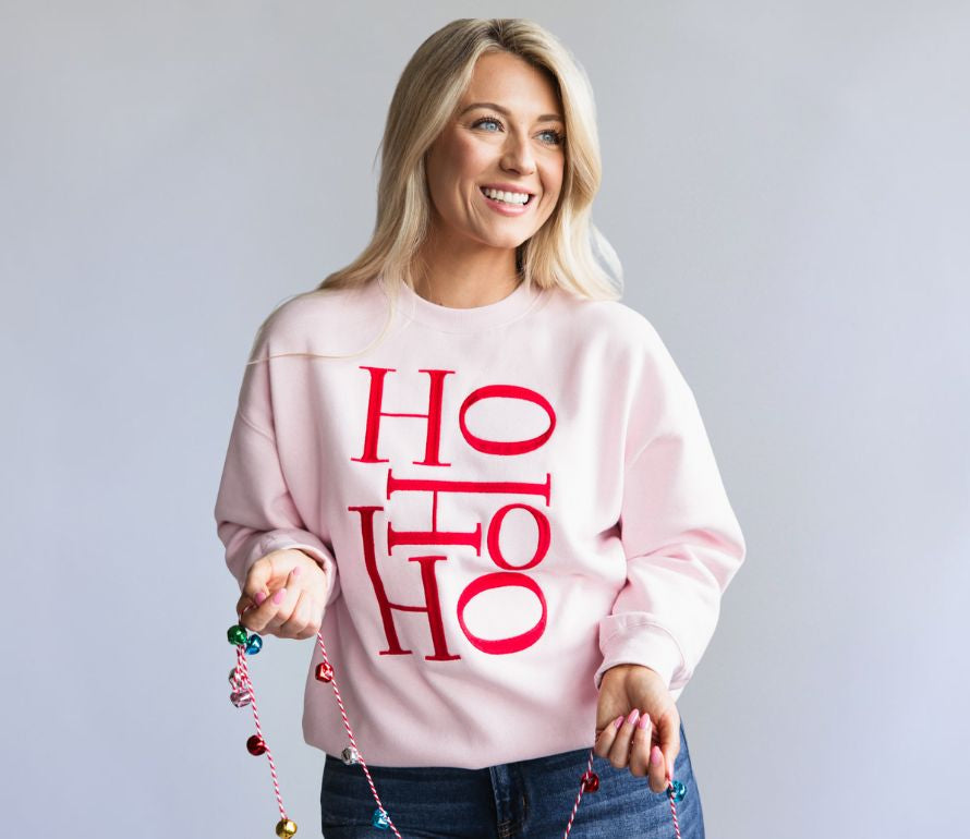 Woman wearing a pink sweatshirt with 'HO HO HO' in red text, holding colorful beads against a light gray background.
