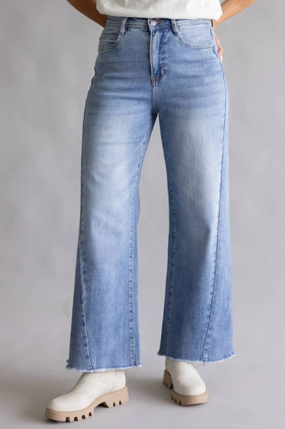 Front view of a model wearing light wash high-rise wide-leg cropped jeans with subtle fading and raw-edge hems, paired with white lug-sole ankle boots against a neutral gray background.